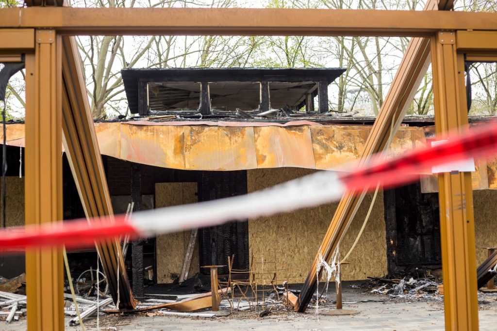 Burnt building with a partially collapsed roof, boarded windows, and charred debris, seen behind a red-and-white caution tape.