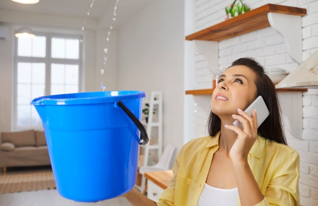 A woman on the phone is carefully holding a blue bucket to catch water leaking from the ceiling; she appears concerned.