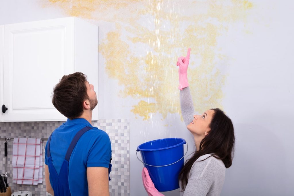 Woman holding a blue bucket under a leaking, water-loss–damaged ceiling while a restoration serviceman inspects the damage.